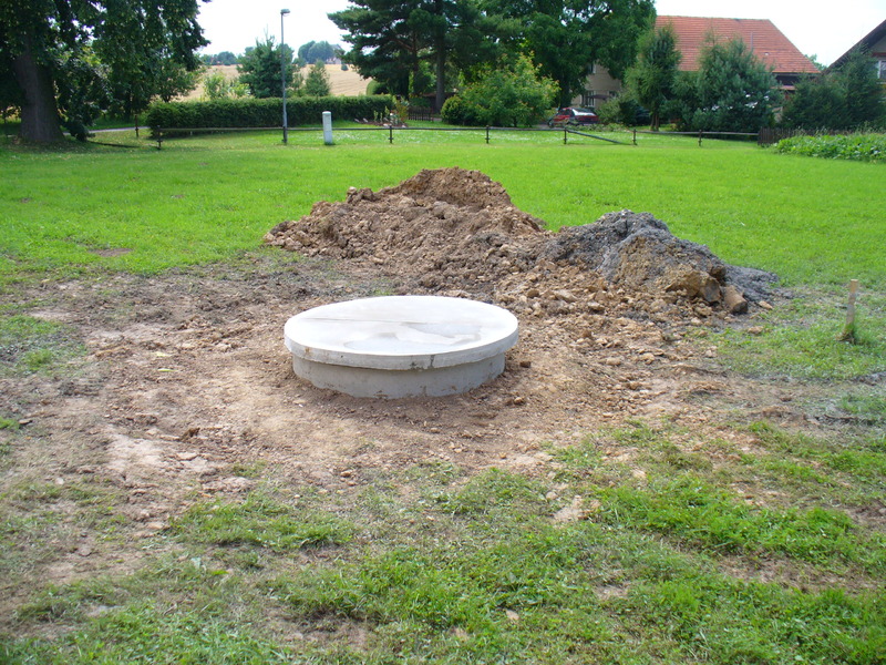 V zemi, uprostřed nedávno rozryté půdy a malých hromádek zeminy, je zasazen betonový poklop kanálu, poblíž hydrogeologických vrtů na travnaté ploše, v pozadí jsou stromy a domy.