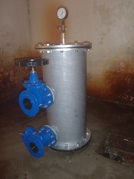 Velké válcové kovové pouzdro filtru pro vrtané studny, s manometrem nahoře a dvěma modrými ventilovými armaturami na boku, umístěné na betonové podlaze u zašpiněné zdi.